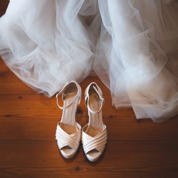 Foto di matrimonio con elegante dettaglio di un abito da sposa in tulle vaporoso che scende delicatamente sopra un pavimento di legno, accompagnato da un paio di scarpe bianche eleganti posizionate di fronte. La scena cattura la bellezza e l’intimità dei preparativi nuziali nel giorno del matrimonio, trasmettendo una sensazione di eleganza e delicatezza. Fotografia di dettaglio wedding che evoca l’emozione e la raffinatezza di un momento speciale.