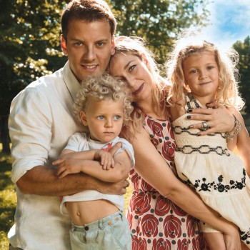 Ritratto di una famiglia composta da madre, padre e due bambini, posata all’aperto in un contesto verde. Il padre abbraccia con affetto il figlio più piccolo, mentre la madre sorride affettuosamente accanto a loro, tenendo la figlia tra le braccia. I bambini hanno espressioni serene e rilassate, mentre la luce naturale illumina i loro volti, evidenziando i capelli biondi dei piccoli. La scena trasmette un senso di unità e calore familiare in una giornata soleggiata. Fotografia di ritratto familiare intima e spontanea.